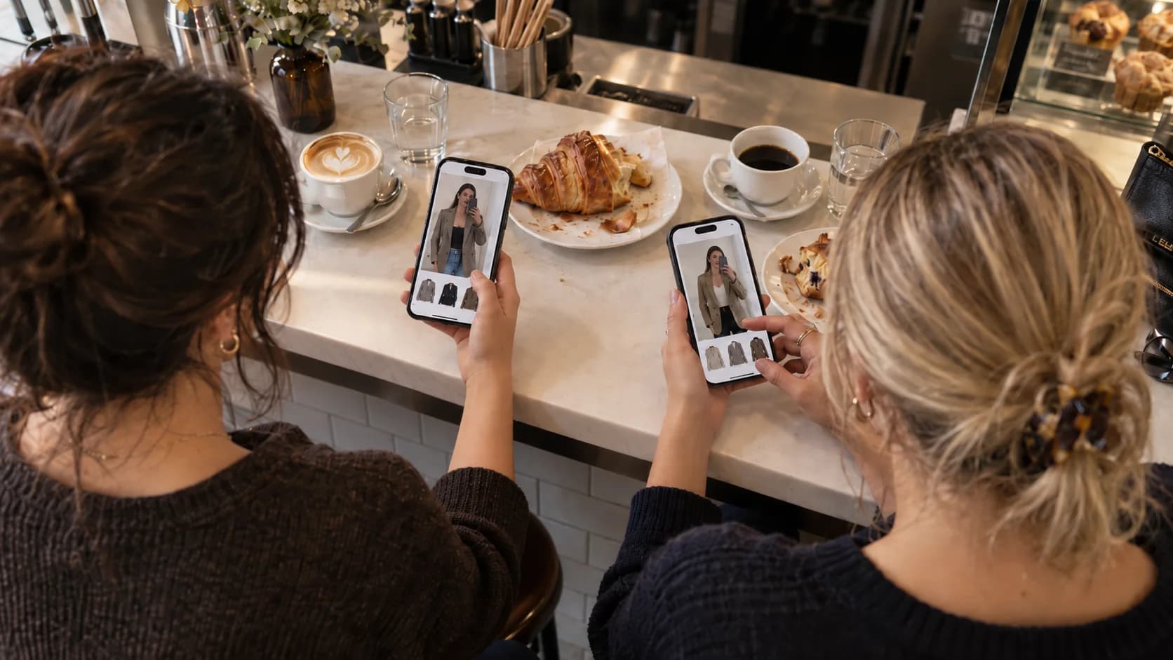 Två kvinnor sitter vid ett cafébord och provar kläder virtuellt på sina mobiler.